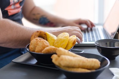 A person typing on a laptop, with a variety of pastries and snacks in black bowls in the foreground. The snacks include empanadas, tequeños, and what appears to be corn arepas. The person has tattoos on their arm and is wearing a casual shirt.