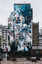 A large, vibrant mural covers the entire side of a building, depicting various people engaged in different activities. The artwork is vivid with a collage-like composition, including scenes of urban life, technology, and historical references. The setting is an urban environment with surrounding buildings and a pedestrian area in front.