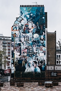 A large, vibrant mural covers the entire side of a building, depicting various people engaged in different activities. The artwork is vivid with a collage-like composition, including scenes of urban life, technology, and historical references. The setting is an urban environment with surrounding buildings and a pedestrian area in front.