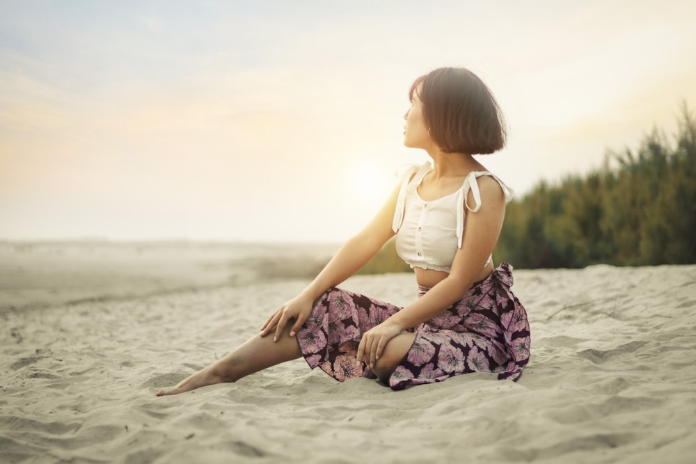 girl in white tank top and purple and white floral skirt sitting on sand during daytime