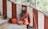 A person with long hair and a beard sits on stone steps, wearing an orange robe and holding a metal bowl. Red and white striped walls are in the background, along with a walking stick leaning against the wall. There is a sense of calm and introspection in a natural, outdoor setting.