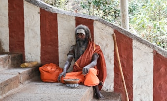 A person with long hair and a beard sits on stone steps, wearing an orange robe and holding a metal bowl. Red and white striped walls are in the background, along with a walking stick leaning against the wall. There is a sense of calm and introspection in a natural, outdoor setting.