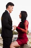 A couple is depicted in an intimate, romantic setting. The woman is wearing an elegant red lace dress, while the man is dressed in a formal black suit with a red bow tie. They are holding hands and gazing into each other's eyes, suggesting a deep connection and affection. The background is subdued, with soft lighting that emphasizes the couple.