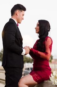 A couple is depicted in an intimate, romantic setting. The woman is wearing an elegant red lace dress, while the man is dressed in a formal black suit with a red bow tie. They are holding hands and gazing into each other's eyes, suggesting a deep connection and affection. The background is subdued, with soft lighting that emphasizes the couple.