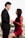 A couple is depicted in an intimate, romantic setting. The woman is wearing an elegant red lace dress, while the man is dressed in a formal black suit with a red bow tie. They are holding hands and gazing into each other's eyes, suggesting a deep connection and affection. The background is subdued, with soft lighting that emphasizes the couple.