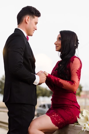 A couple is depicted in an intimate, romantic setting. The woman is wearing an elegant red lace dress, while the man is dressed in a formal black suit with a red bow tie. They are holding hands and gazing into each other's eyes, suggesting a deep connection and affection. The background is subdued, with soft lighting that emphasizes the couple.