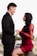A couple is depicted in an intimate, romantic setting. The woman is wearing an elegant red lace dress, while the man is dressed in a formal black suit with a red bow tie. They are holding hands and gazing into each other's eyes, suggesting a deep connection and affection. The background is subdued, with soft lighting that emphasizes the couple.