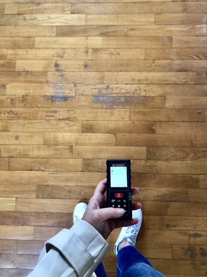 A person holding a handheld device with a screen displaying various information. The floor is made of light brown wooden planks, and the person is wearing white sneakers and a light-colored coat.
