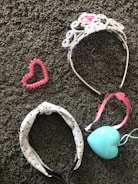 A collection of accessories lies on a textured gray carpet. A sparkling tiara adorned with pink and purple gemstones is beside a pink heart-shaped beaded bracelet. A headband with a white base and black and red pattern is next to a pink-ribboned tiara-like headband and a light blue heart-shaped pendant or ornament.