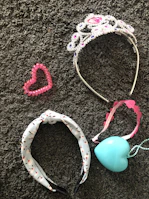 A collection of accessories lies on a textured gray carpet. A sparkling tiara adorned with pink and purple gemstones is beside a pink heart-shaped beaded bracelet. A headband with a white base and black and red pattern is next to a pink-ribboned tiara-like headband and a light blue heart-shaped pendant or ornament.