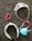 A collection of accessories lies on a textured gray carpet. A sparkling tiara adorned with pink and purple gemstones is beside a pink heart-shaped beaded bracelet. A headband with a white base and black and red pattern is next to a pink-ribboned tiara-like headband and a light blue heart-shaped pendant or ornament.