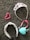 A collection of accessories lies on a textured gray carpet. A sparkling tiara adorned with pink and purple gemstones is beside a pink heart-shaped beaded bracelet. A headband with a white base and black and red pattern is next to a pink-ribboned tiara-like headband and a light blue heart-shaped pendant or ornament.