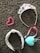 A collection of accessories lies on a textured gray carpet. A sparkling tiara adorned with pink and purple gemstones is beside a pink heart-shaped beaded bracelet. A headband with a white base and black and red pattern is next to a pink-ribboned tiara-like headband and a light blue heart-shaped pendant or ornament.