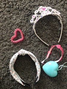 A collection of accessories lies on a textured gray carpet. A sparkling tiara adorned with pink and purple gemstones is beside a pink heart-shaped beaded bracelet. A headband with a white base and black and red pattern is next to a pink-ribboned tiara-like headband and a light blue heart-shaped pendant or ornament.