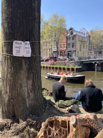A tree with signs and QR codes is in the foreground near a canal. People are seated by the water's edge, some using their phones. A boat with passengers wearing orange life jackets is passing by. In the background, there are colorful and traditional buildings typical of Amsterdam's architecture.