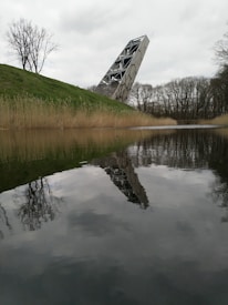 A modern, angular structure tilts dramatically, reflected in a calm body of water. Bare trees and dry reeds line the water's edge, contrasting with the structure's sleek design. Overcast skies contribute to the serene yet enigmatic atmosphere.