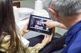 A person is sitting in a dental office looking at a dental X-ray on a tablet device held by another person. The individual in the foreground is pointing at the images on the screen. Dental equipment and a sink are visible in the background.