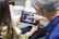 A person is sitting in a dental office looking at a dental X-ray on a tablet device held by another person. The individual in the foreground is pointing at the images on the screen. Dental equipment and a sink are visible in the background.