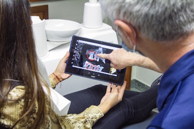 A person is sitting in a dental office looking at a dental X-ray on a tablet device held by another person. The individual in the foreground is pointing at the images on the screen. Dental equipment and a sink are visible in the background.