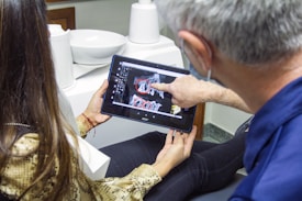 A person is sitting in a dental office looking at a dental X-ray on a tablet device held by another person. The individual in the foreground is pointing at the images on the screen. Dental equipment and a sink are visible in the background.