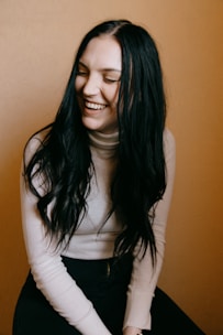 A person with long dark hair is laughing joyfully while looking slightly down. They are wearing a light-colored turtleneck sweater and sitting against a warm, mustard-colored background.