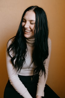 A person with long dark hair is laughing joyfully while looking slightly down. They are wearing a light-colored turtleneck sweater and sitting against a warm, mustard-colored background.