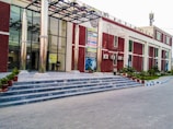 A building with a modern design featuring a glass facade and tall metal columns at the entrance. It has a series of steps leading up to the entrance, where potted plants are placed on either side. Several posters are displayed on the front wall, and there are air conditioning units attached to the building’s exterior. The pathway in front is paved, and there are green plants and a neatly maintained lawn on the surrounding grounds.