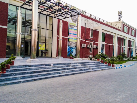A building with a modern design featuring a glass facade and tall metal columns at the entrance. It has a series of steps leading up to the entrance, where potted plants are placed on either side. Several posters are displayed on the front wall, and there are air conditioning units attached to the building’s exterior. The pathway in front is paved, and there are green plants and a neatly maintained lawn on the surrounding grounds.
