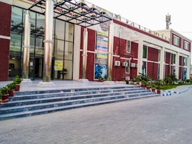 A building with a modern design featuring a glass facade and tall metal columns at the entrance. It has a series of steps leading up to the entrance, where potted plants are placed on either side. Several posters are displayed on the front wall, and there are air conditioning units attached to the building&rsquo;s exterior. The pathway in front is paved, and there are green plants and a neatly maintained lawn on the surrounding grounds.