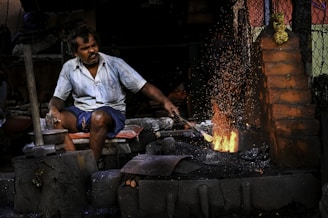 A man works intensely at a blacksmith's forge, surrounded by tools. He is wearing a light blue shirt and dark shorts, focusing on the task of hammering metal. Bright orange sparks fly up from the glowing furnace, illuminating the dimly lit workshop filled with bricks and metal objects.