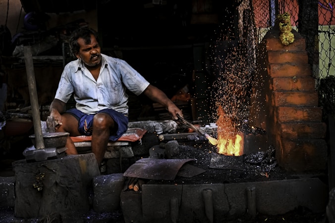 A man works intensely at a blacksmith's forge, surrounded by tools. He is wearing a light blue shirt and dark shorts, focusing on the task of hammering metal. Bright orange sparks fly up from the glowing furnace, illuminating the dimly lit workshop filled with bricks and metal objects.