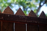 A close-up view of a wooden fence with pointed tops. The wood appears aged and textured, with visible grain and imperfections. In the background, there is a blur of green foliage from trees, suggesting a natural setting.