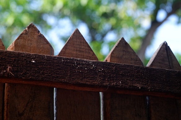 A close-up view of a wooden fence with pointed tops. The wood appears aged and textured, with visible grain and imperfections. In the background, there is a blur of green foliage from trees, suggesting a natural setting.