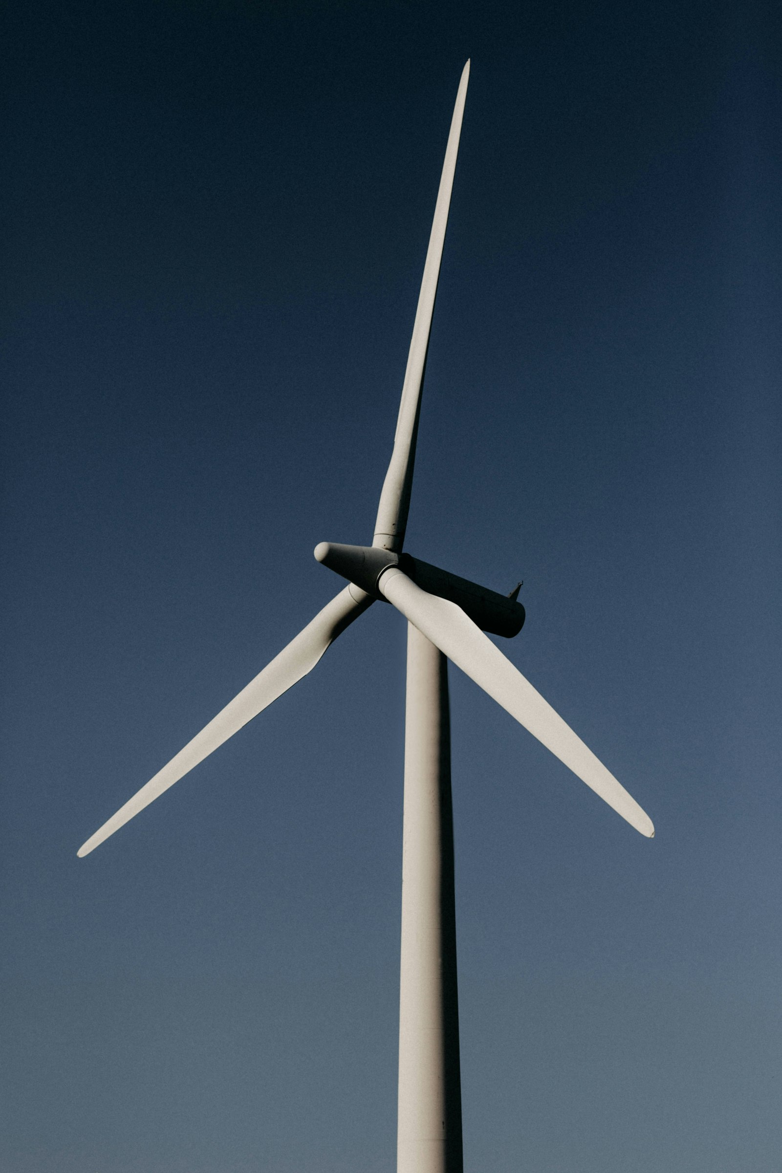 A tall white wind turbine tower rising against a clear blue sky, seen from the base looking up toward the nacelle