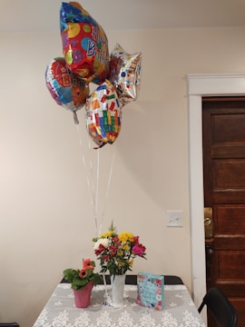A small table is adorned with a floral-patterned tablecloth. On it, there are two plant arrangements; one is a bouquet of mixed flowers in a white vase, and the other is a pink potted plant. A card with floral designs and a 'Happy Birthday' theme is placed on the table. Above the table, a cluster of colorful 'Happy Birthday' balloons are floating, tied to the table.