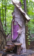 A whimsical wooden structure resembling a small house is carved into the trunk of a tree. The door is painted purple with ornate black designs and an arched window at the top. Steps made from slices of wood lead up to the door. The background is a lush green forest, creating an enchanting and fairy-tale-like ambiance.