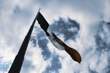 A Mexican flag waves on a tall flagpole against a backdrop of a partly cloudy sky. The perspective is from below, emphasizing the height of the pole and the wide expanse of the sky.