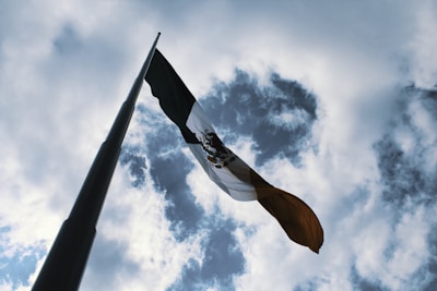 A Mexican flag waves on a tall flagpole against a backdrop of a partly cloudy sky. The perspective is from below, emphasizing the height of the pole and the wide expanse of the sky.