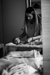 A woman with long hair is gently taking care of a baby on a changing table. The baby is lying on a patterned cover and looking towards the camera. The scene is captured in black and white, giving it a serene and classic feel. There are soft fabrics and towels in the foreground, adding to the nurturing atmosphere.