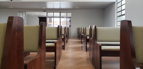 A room with rows of empty wooden chairs with cushioned seats, arranged neatly in a symmetrical pattern. The space has large windows on one side allowing natural light to fill the room, and the flooring is polished wood.