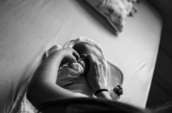 A black and white image of a person gently swaddling or dressing a baby lying on a bed. The scene captures an intimate and tender moment. Soft lighting and a focus on hands create a calm atmosphere.