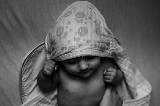 A black and white photograph of a baby with eyes closed, partially covered by a hooded towel. The baby appears to be calm and content, with small fists slightly clenched near their face. The towel features a simple pattern.