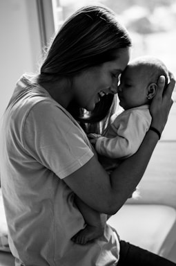 A woman gently cradles a baby close to her face, both appearing to share a tender moment. The scene is black and white, highlighting the emotional connection between them as they touch their foreheads together.