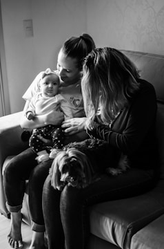 A tender moment is captured in black and white, featuring two adults seated on a couch with an infant held by one of them. The baby is dressed in a light-colored outfit with a headband, looking towards the camera. Next to the adults, a small dog sits on the lap of one person, seemingly relaxed. The overall setting appears intimate and familial.