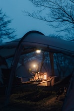 A camping setup under a tent with a picnic table and chairs. On the table, there are lanterns providing a warm light amidst the cool, dim evening or dawn ambiance. Shadows and outlines of trees are visible in the background, suggesting a serene, natural setting.