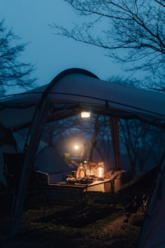 A camping setup under a tent with a picnic table and chairs. On the table, there are lanterns providing a warm light amidst the cool, dim evening or dawn ambiance. Shadows and outlines of trees are visible in the background, suggesting a serene, natural setting.