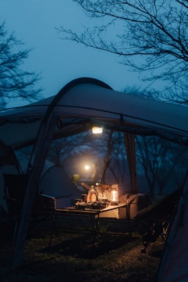 A camping setup under a tent with a picnic table and chairs. On the table, there are lanterns providing a warm light amidst the cool, dim evening or dawn ambiance. Shadows and outlines of trees are visible in the background, suggesting a serene, natural setting.