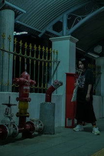 A person stands next to a red fire hydrant with metallic pipes attached. There is a yellow and black decorative metal fence in the background, along with concrete pillars and an arched roof structure above.