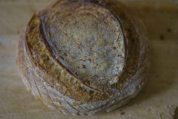 A round loaf of artisan bread with a crispy, textured crust and a deep brown color rests on a wooden surface. The bread's surface features a pattern of concentric cracks and lines, highlighting its rustic quality and artisanal craftsmanship.