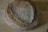 A round loaf of artisan bread with a crispy, textured crust and a deep brown color rests on a wooden surface. The bread's surface features a pattern of concentric cracks and lines, highlighting its rustic quality and artisanal craftsmanship.